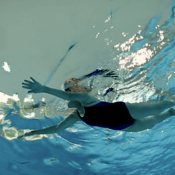 Still from Eskape. Underwater shot of a woman in a one-piece swimsuit and swim cap, swimming with arms outstretched in front of her. The water is bright blue and in areas appears silver from the reflection of light on the surface.