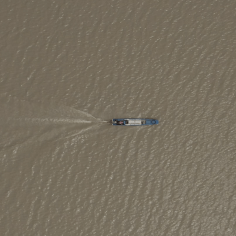 Still from Eskape. Overhead bird-eye view of a long blue and white motorized boat. The boat is centered in the middle of the frame, and is moving to the right, cutting across a wide expanse of water and leaving a wake behind it. The water is light brown in color, and the sun reflects off the waves.