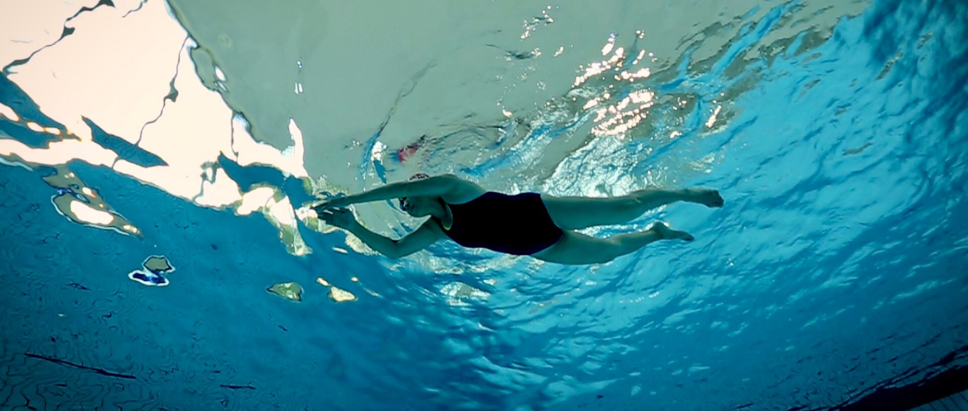 Still from Eskape. Underwater shot of a woman in a one-piece swimsuit and swim cap, swimming with arms crossing in front of her. The water is bright blue and in areas appears silver from the reflection of light on the surface.