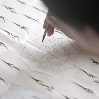Still from Hidden Letters. Close up of a woman's hand drawing a floral design in ink on paper. Around the floral design, there are vertical rows of "Nushu" script.