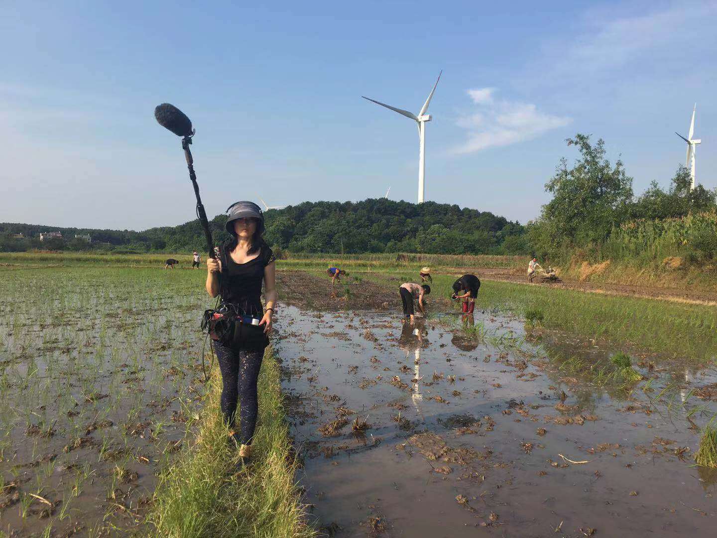 Production still from Hidden Letters. A woman is walking on an elevated grassy row in a rice field. She is wearing headphones and a utility bag around her waist, and is carrying a boom microphone. Behind her, there are several women bent over working in the rice field, and two white windmills in the distance.