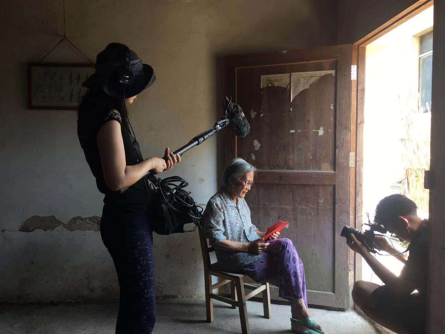 Production still from Hidden Letters. An elderly woman is sitting on a chair, looking at a piece of red paper that she holds in her hand. She is illuminated by light coming in through the doorway. To her left, a woman is holding a boom mic, and to her front, a man is crouched with a camera pointed in her direction.