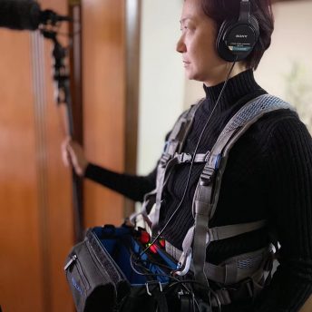 Production still from Hidden Letters. Co-director Qing Zhao is pictured from the waist up. She is wearing a black turtleneck and a grey harness and a camera bag attached with cables. She has on headphones over her ears, and is holding a boom microphone in her right hand.