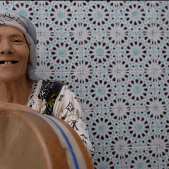 Still from Machtat. A woman in a headwrap smiles at the camera, while she plays a drum.