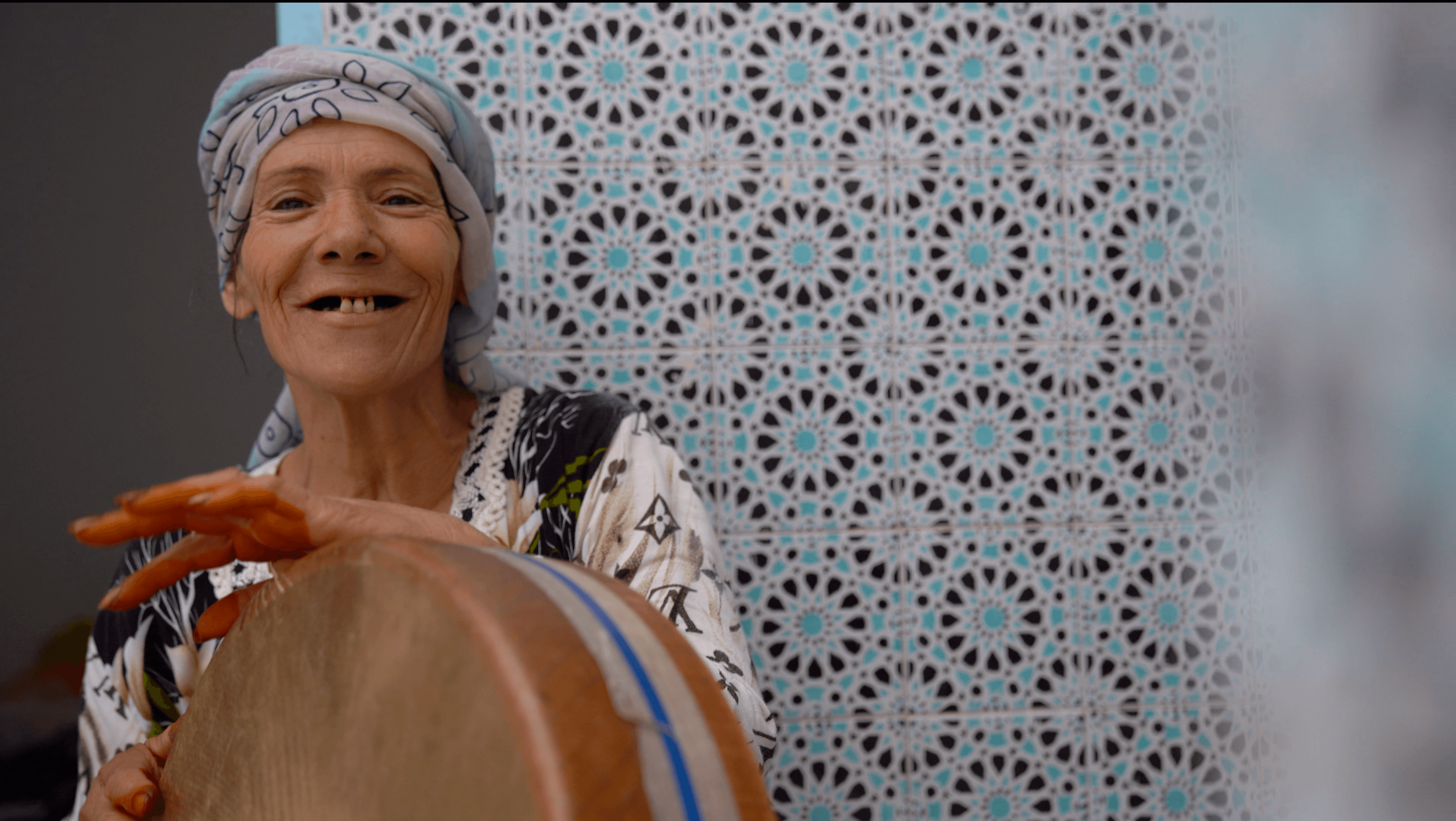 Still from Machtat. A woman in a headwrap smiles at the camera, while she plays a drum.