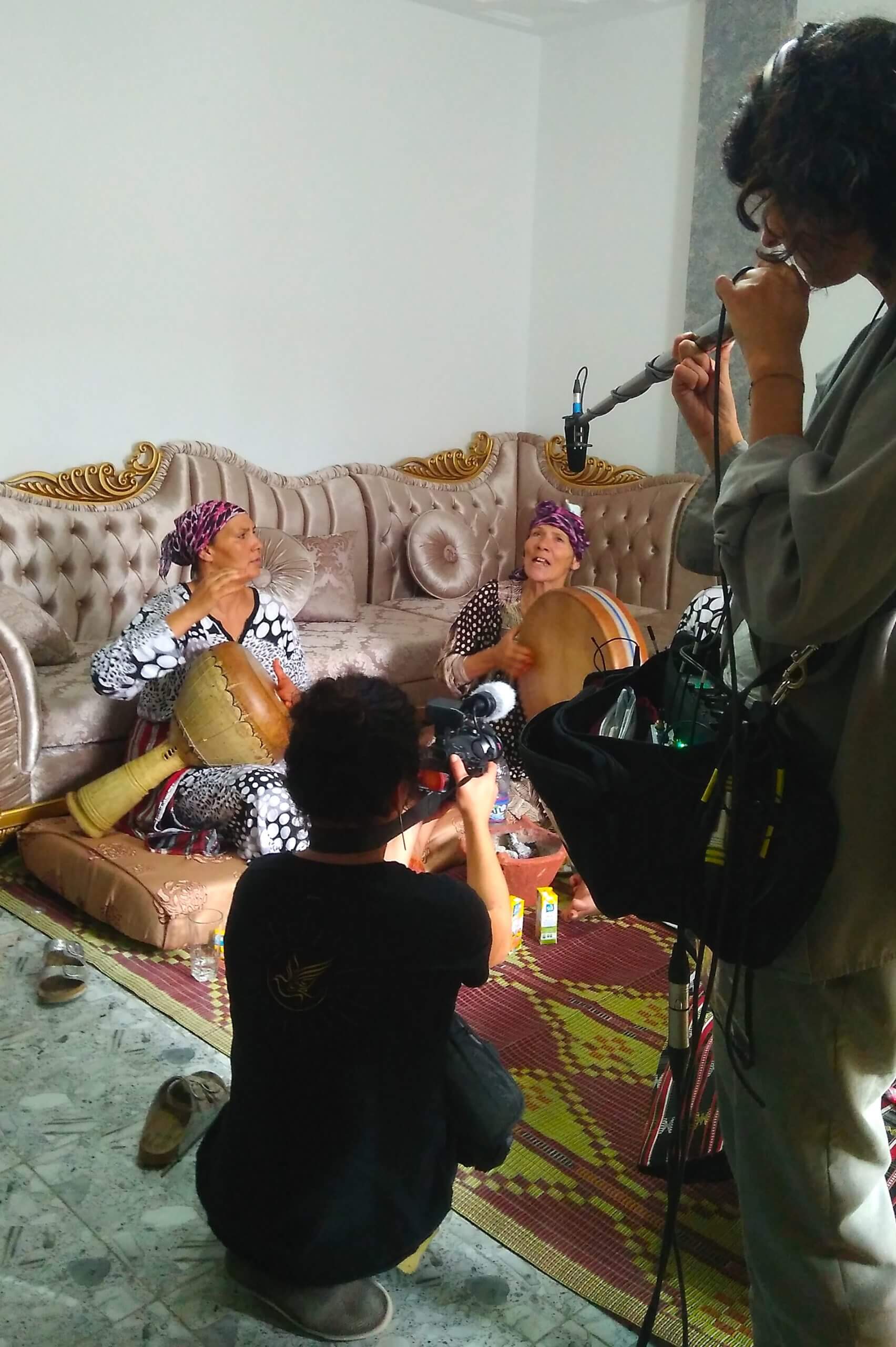 Production still from Machtat. Two film crew members, one with a camera and one with a boom mic stand in front of two women in headwraps. They are sitting on a coushion on the floor in front of a couch playing drums and singing.