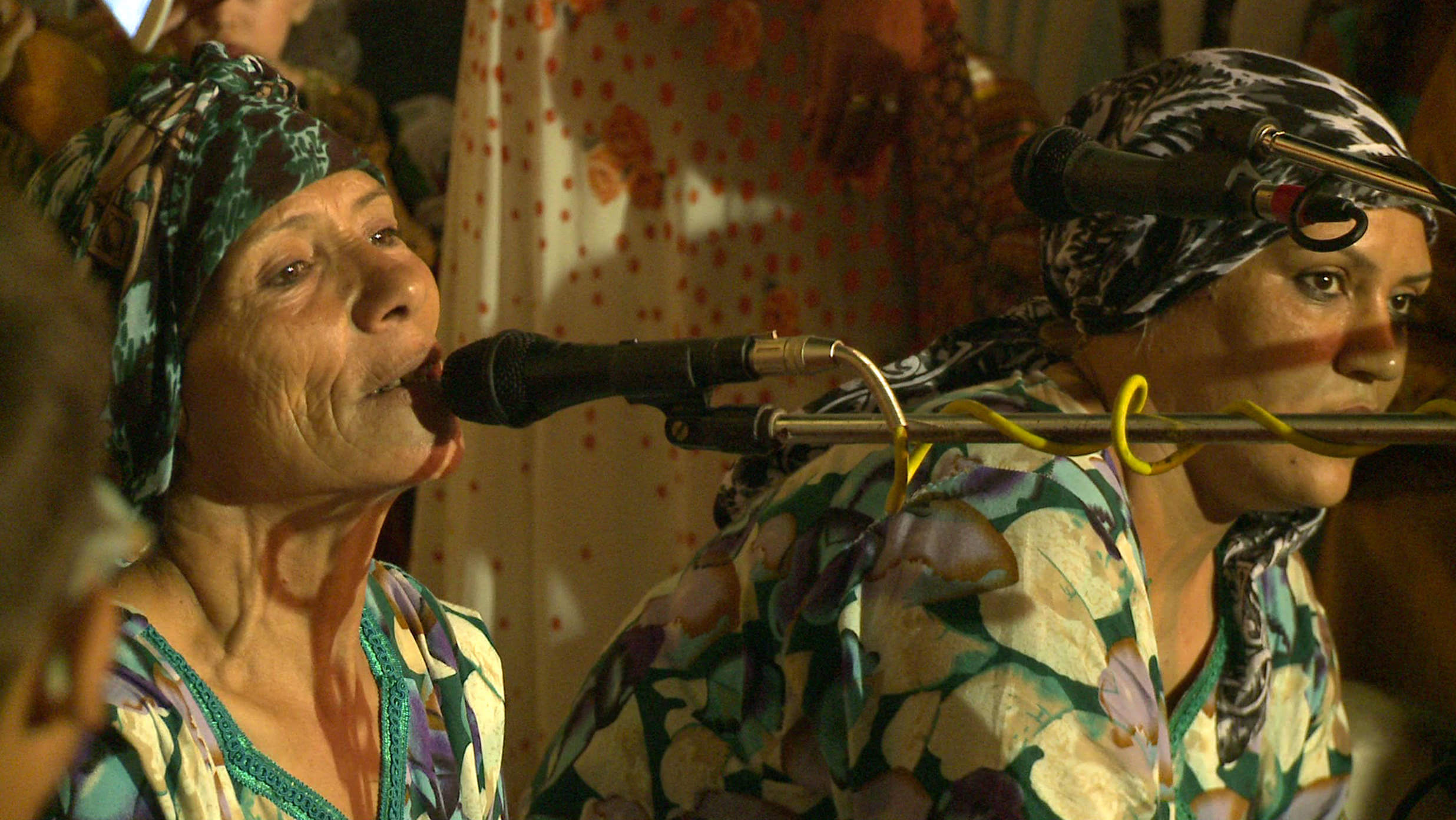 Still from Machtat. Two women in colorful dresses and headwraps. One is singing with a microphone.