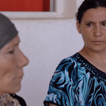 Still from Machtat. One woman looks to another woman in a headwrap who is closer to the camera and slightly out of focus.