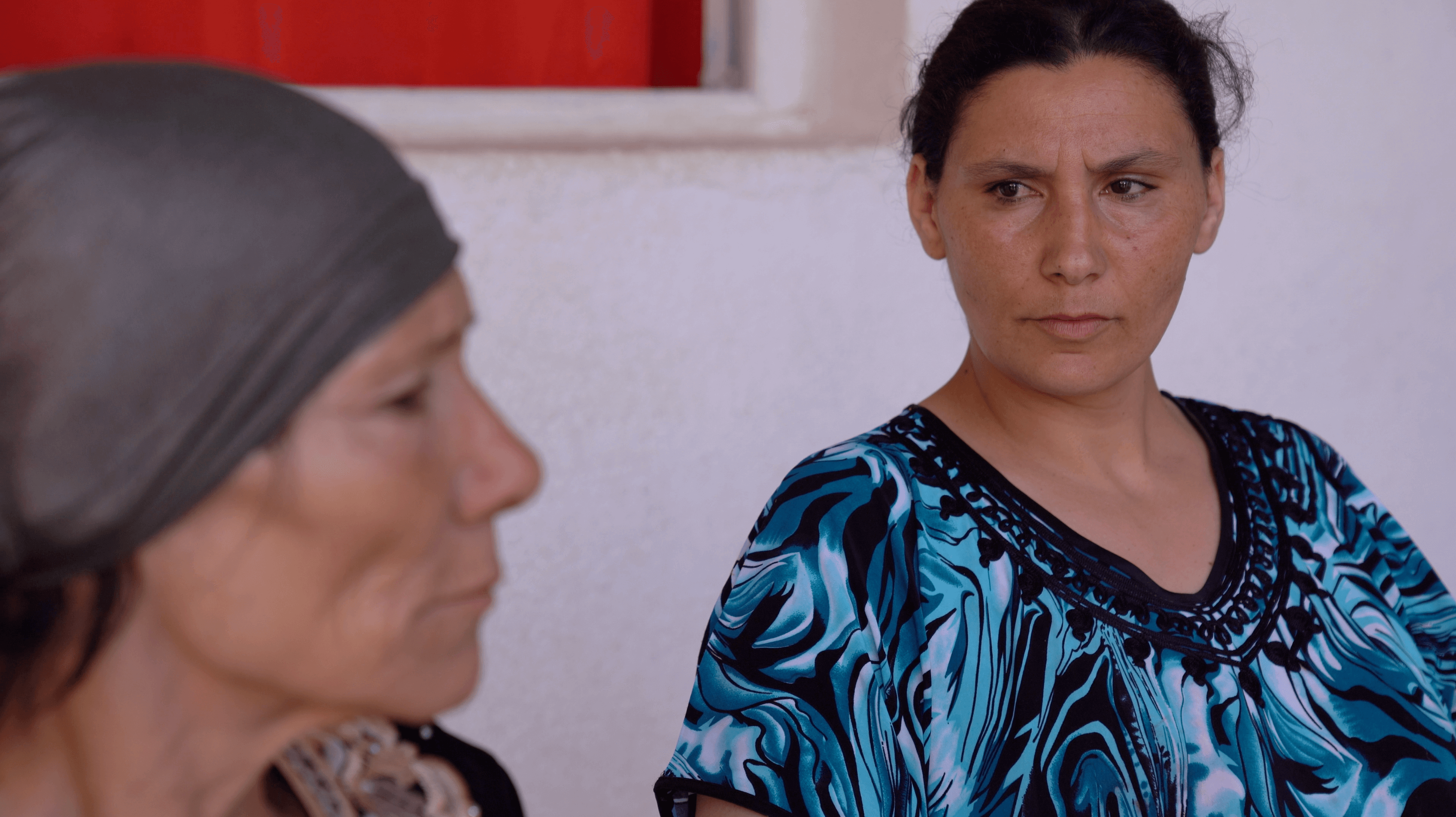 Still from Machtat. One woman looks to another woman in a headwrap who is closer to the camera and slightly out of focus.
