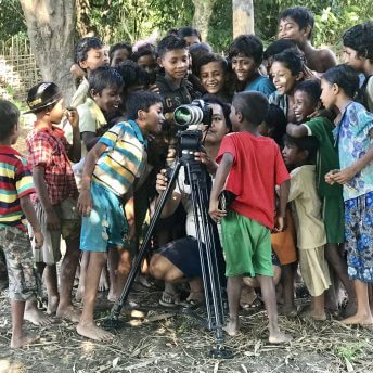 Production still from Midwives. Twenty kids are surrounding a camera man with a camera on a tripod, looking at the footage.