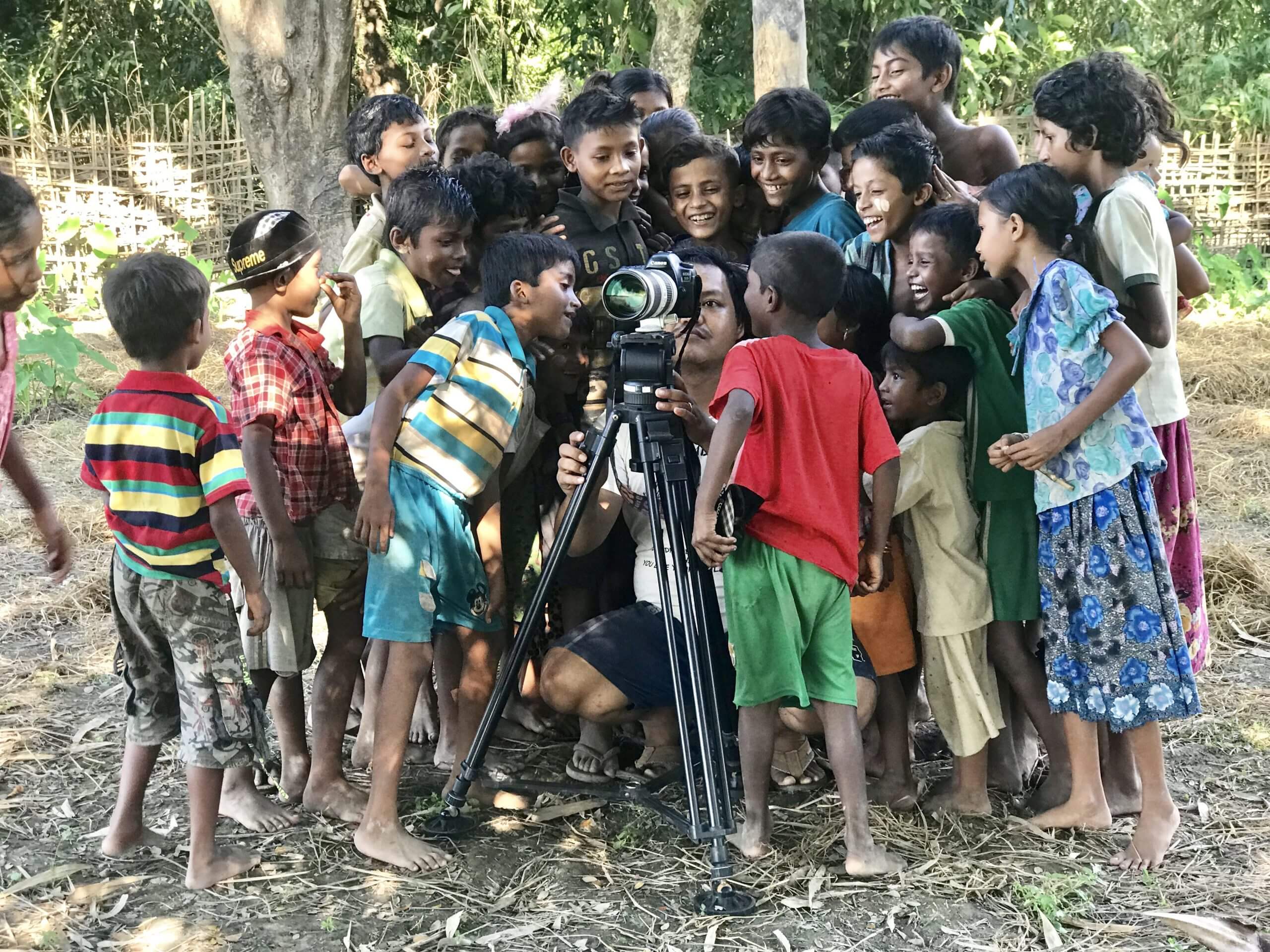 Production still from Midwives. Twenty kids are surrounding a camera man with a camera on a tripod, looking at the footage.
