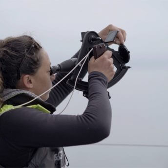 A woman in a long sleeve shirt, puffy vest, and sunglasses looking through a sextant out at a gray horizon in a body of water