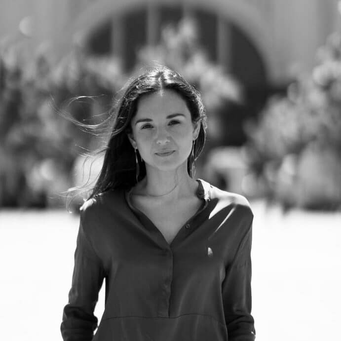 Black and white headshot of Ainara Vera from the waist up wearing a long sleeve dark blouse standing in front of a blurred building and trees.