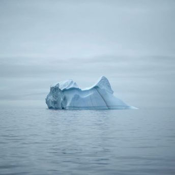 An iceberg in the middle of a body of water on the horizon against cloudy grey skies
