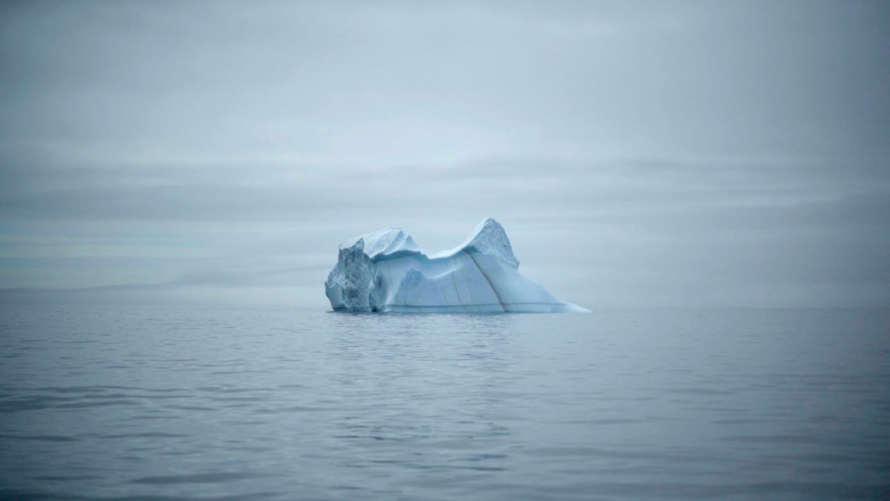 An iceberg in the middle of a body of water on the horizon against cloudy grey skies
