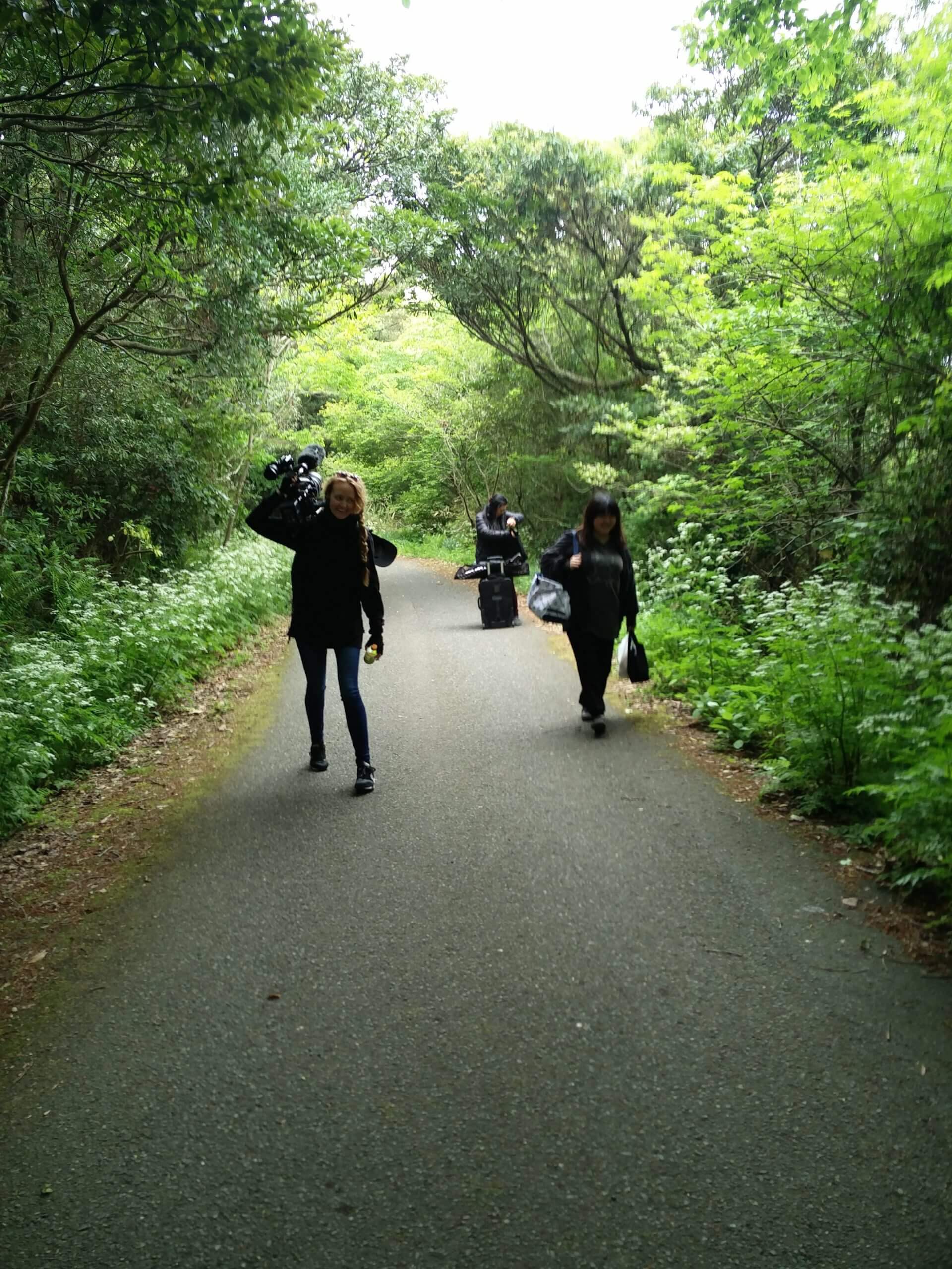 Production still from All of Our Heartbeats Are Connected Through Exploding Stars. Three people from the crew are walking in a forest path, they are carrying equipment.