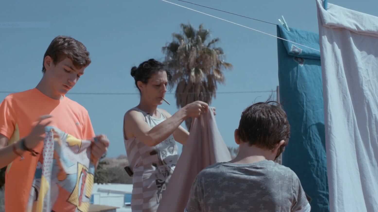 A still from The Boy and the Suit of Lights. A woman smoking a cigarette holds up a large piece of fabric. She stands in front of a palm tree and a little boy is in front of her, his back facing the camera.