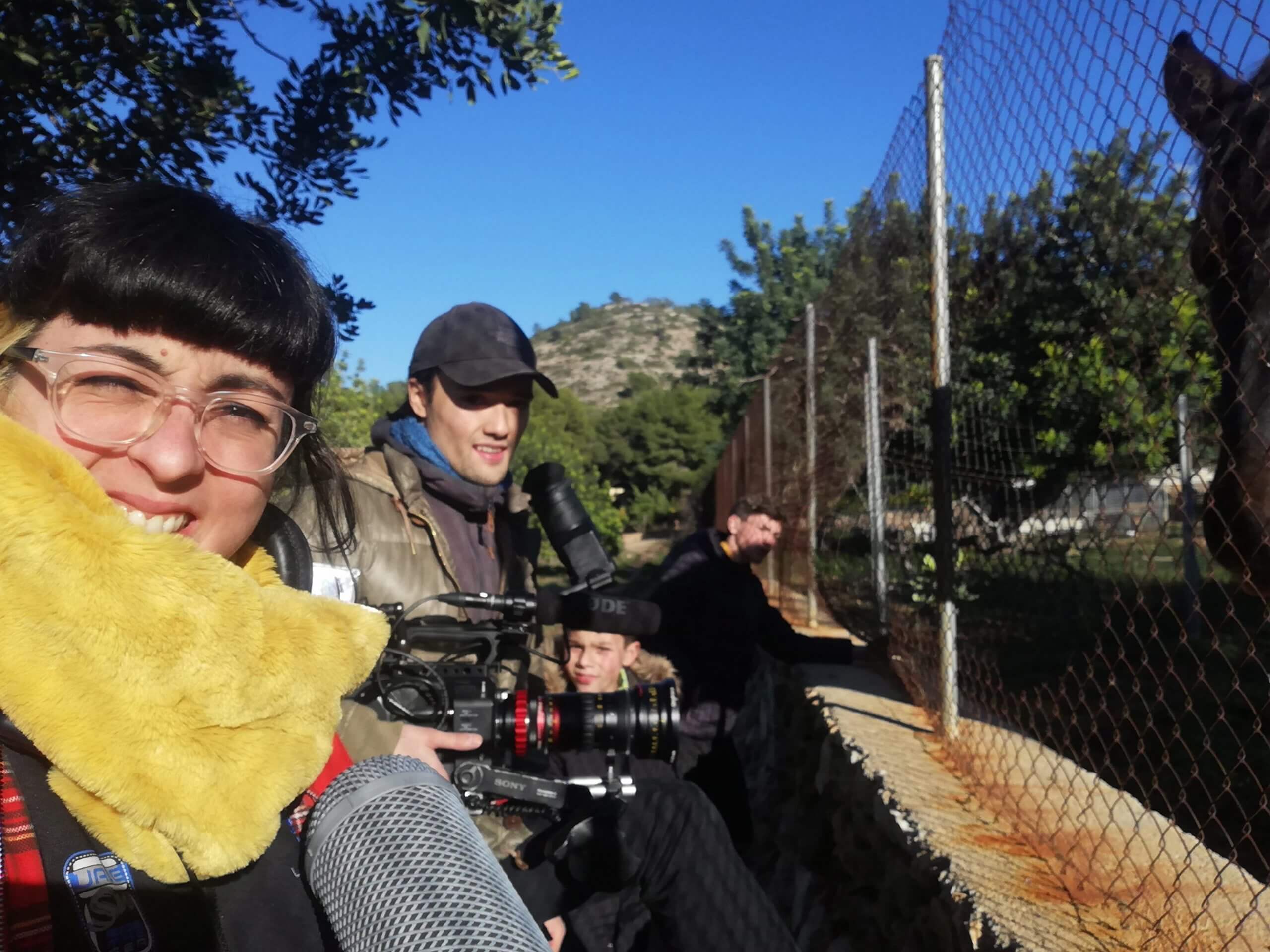 A production still from The Boy and the Suit of Lights. Inma de Reyes takes a selfie of herself and two crew members in front of a fence. One of the crew members holds a large camera.