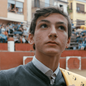 A still from The Boy and the Suit of Lights. A close-up shot of a young boy wearing a grey suit and a yellow cloth draped over his left shoulder. Behind him, a crowd of people sits in stadium seating. He has short brown hair and brown eyes.