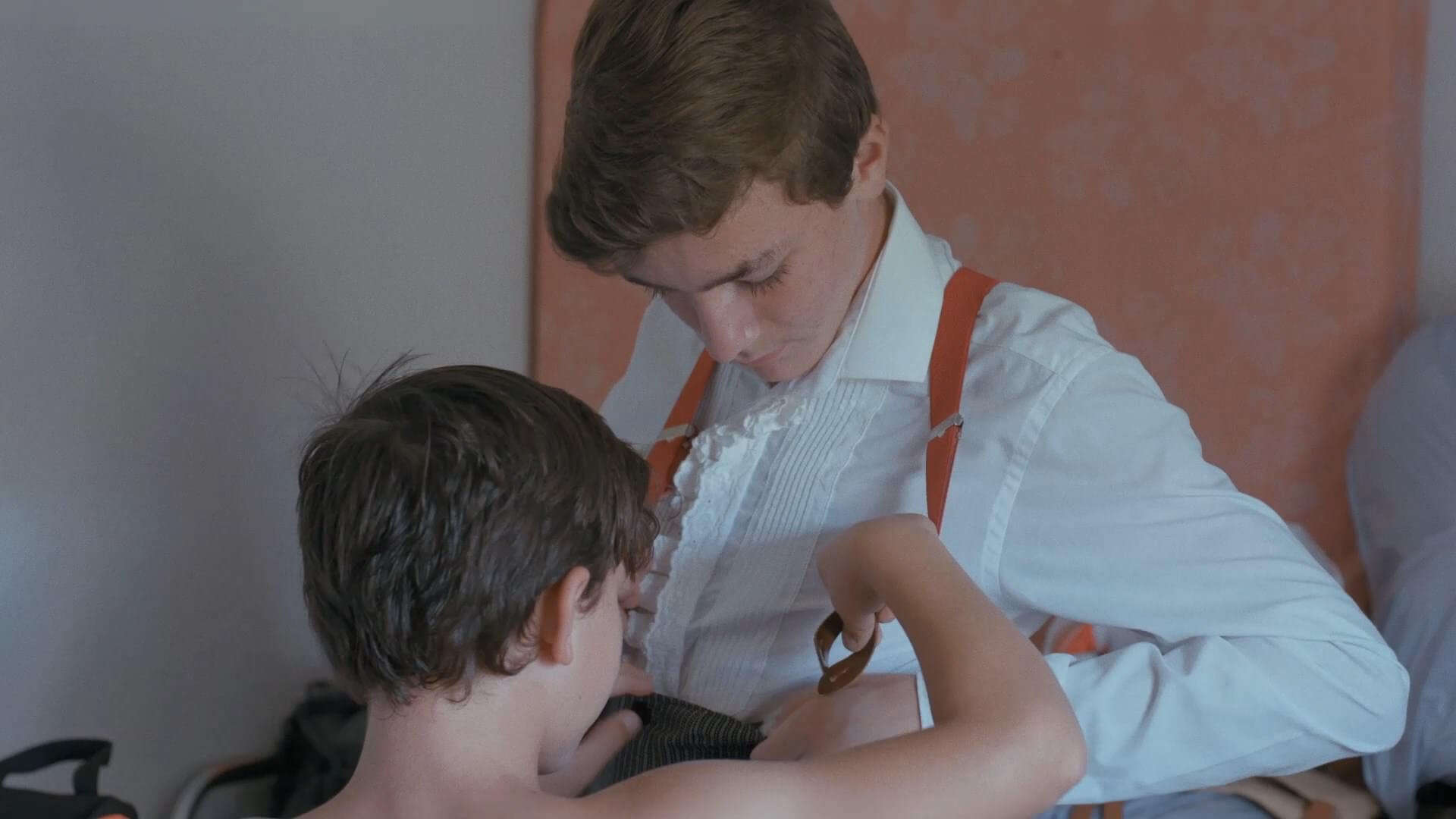 A still from The Boy and the Suit of Lights. One boy helps another fasten his red suspender to his pants. The boy in the suspenders wears a white button-down shirt underneath.