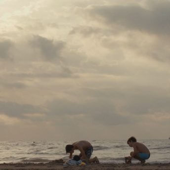 A still from The Boy and the Suit of Lights. Two young boys crouch down on the beach. The ocean and a cloudy sky are in the background in various shades of white, grey, and pale blue.