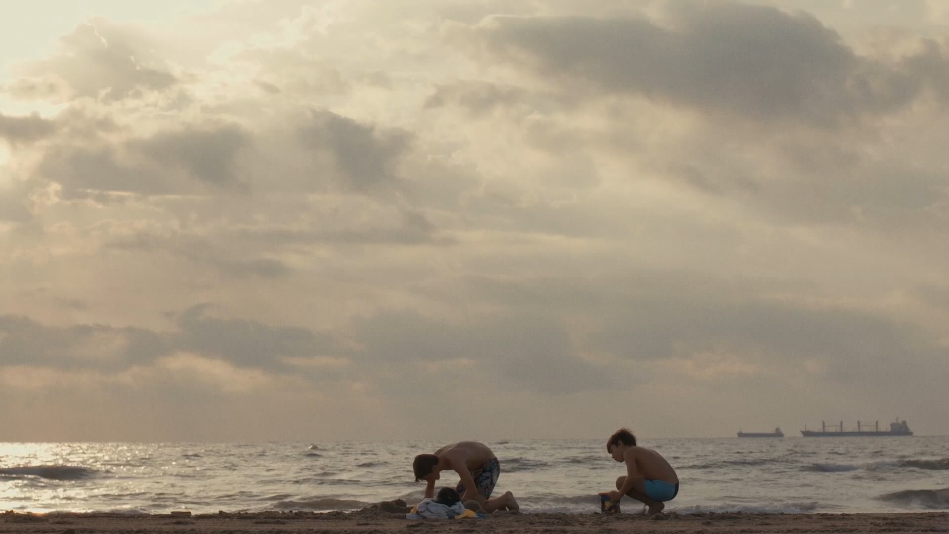 A still from The Boy and the Suit of Lights. Two young boys crouch down on the beach. The ocean and a cloudy sky are in the background in various shades of white, grey, and pale blue.