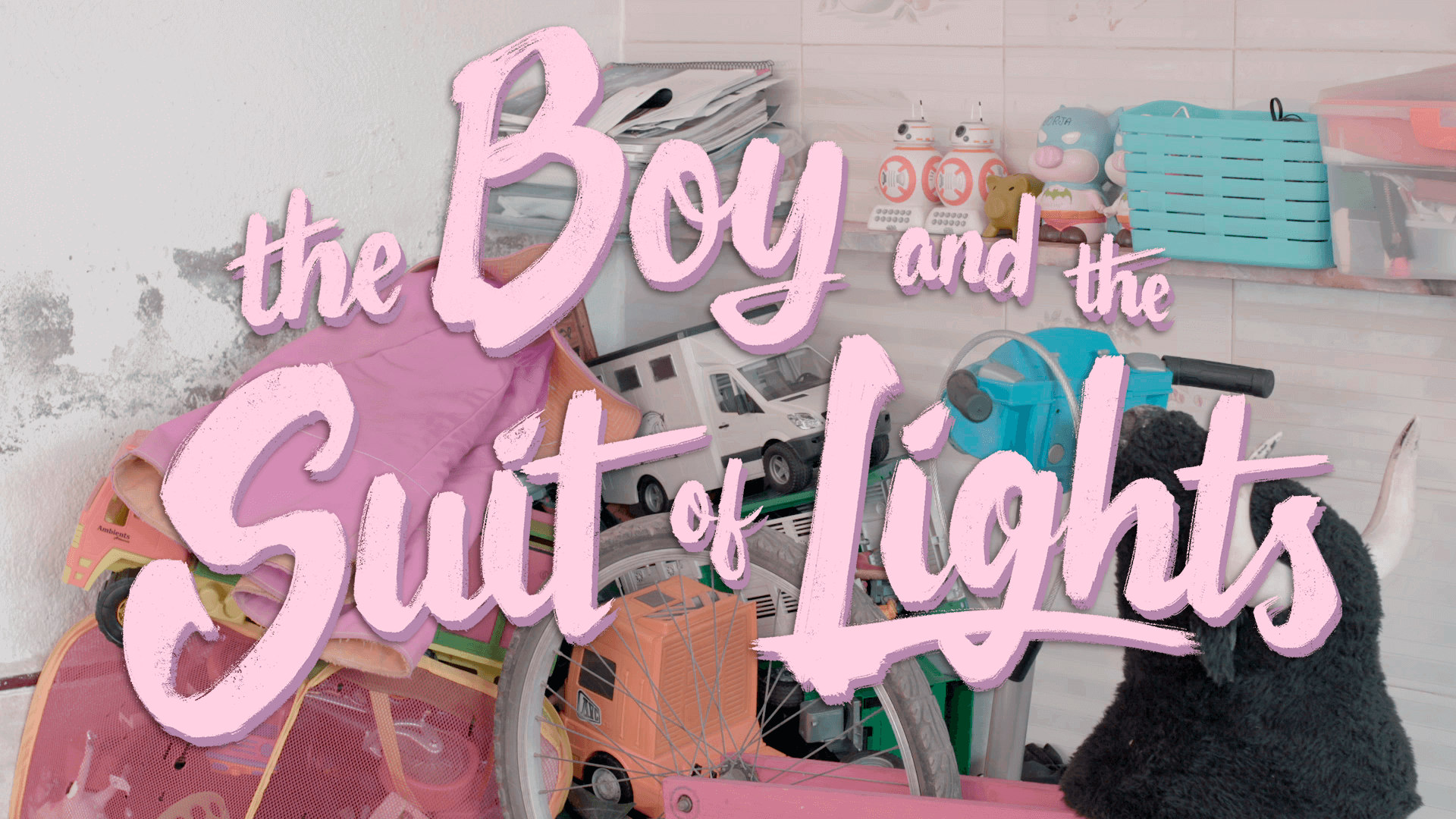 A photo of the graphic for The Boy and the Suit of Lights. The title is spelled out in pink cursive font over a pile of toys sitting in a white room.