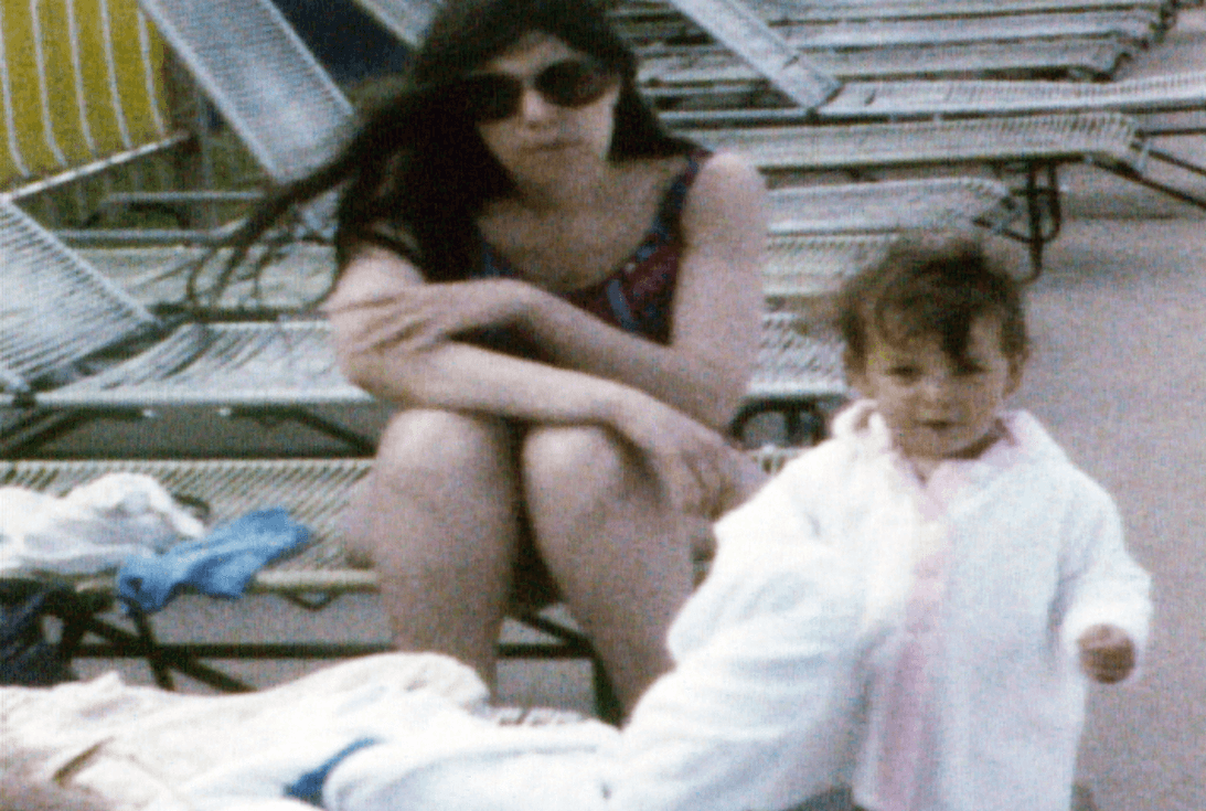 Photography of a woman sitting on a pool chair. In front of her is a toddler with a big white towel; the toddler is looking at the camera.