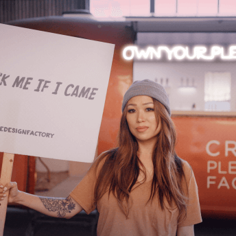 A still from The Dilemma of Desire. A person with long hair holds a sign that reads, "ask me if I came". They are wearing a pink t-shirt and grey beanie.