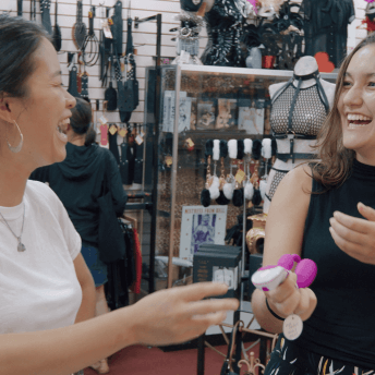 A Still from The Dilemma of Desire. Two people stand in a sex shop, holding sex toys in their hands. They are laughing.