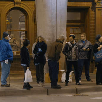 A still from the Dilemma of Desire. A line of people standing on the sidewalk, outside of a building.
