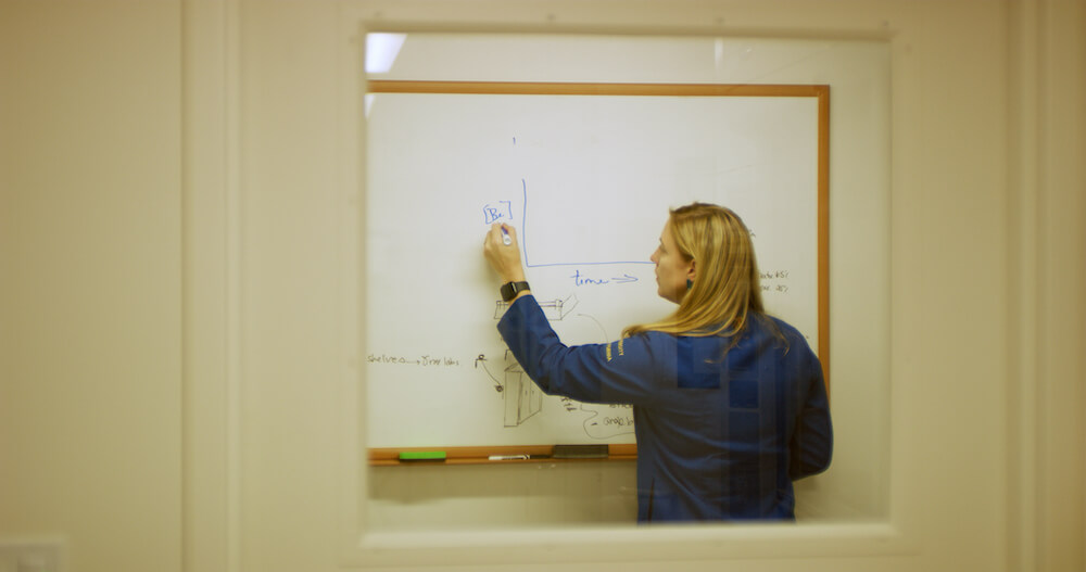 View through a window of a woman in a blue shirt writing labels of a graph onto a whiteboard.