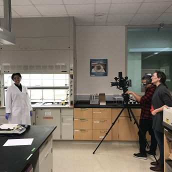 Two people behind a tripod with a camera in a science lab, filming a smiling woman in a white lab coat