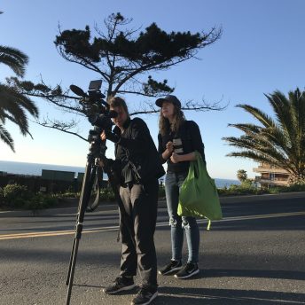 Two people outside with a camera on a tripod in the middle of a paved street, with one person filming while the other watches