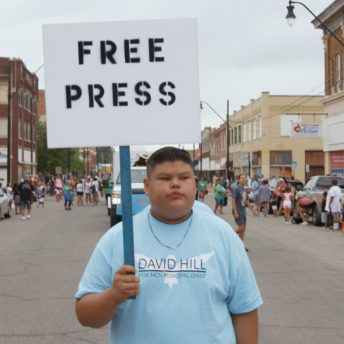 An Indigenous teen walks in the 2019 Muscogee Festival parade in Okmulgee, OK. He is wearing a light blue campaign shirt and holding a sign with the words "Free Press."