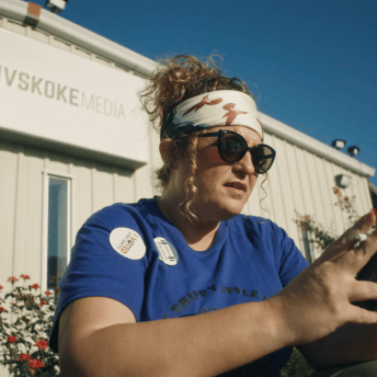 Indigenous journalist Angel Ellis sits in front of Mvskoke Media wearing a bandana , sunglasses and a "I Voted" sticker on her blue T-shirt. She's smoking a cigarette while scrolling through her cell phone on a warm summer evening.