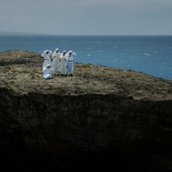 Still from Landfall. Wide shot of a group of nuns in white habits on top of a rock in the middle of a wide expanse of water. Several of the nuns are holding onto their habits due to the wind, and white waves from the wind dot the expanse of water.