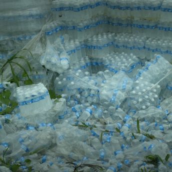 Still from Landfall. Hundreds of plastic bottles are packaged in plastic. Some of the plastic is broken open and the bottles have been spread all over the ground.