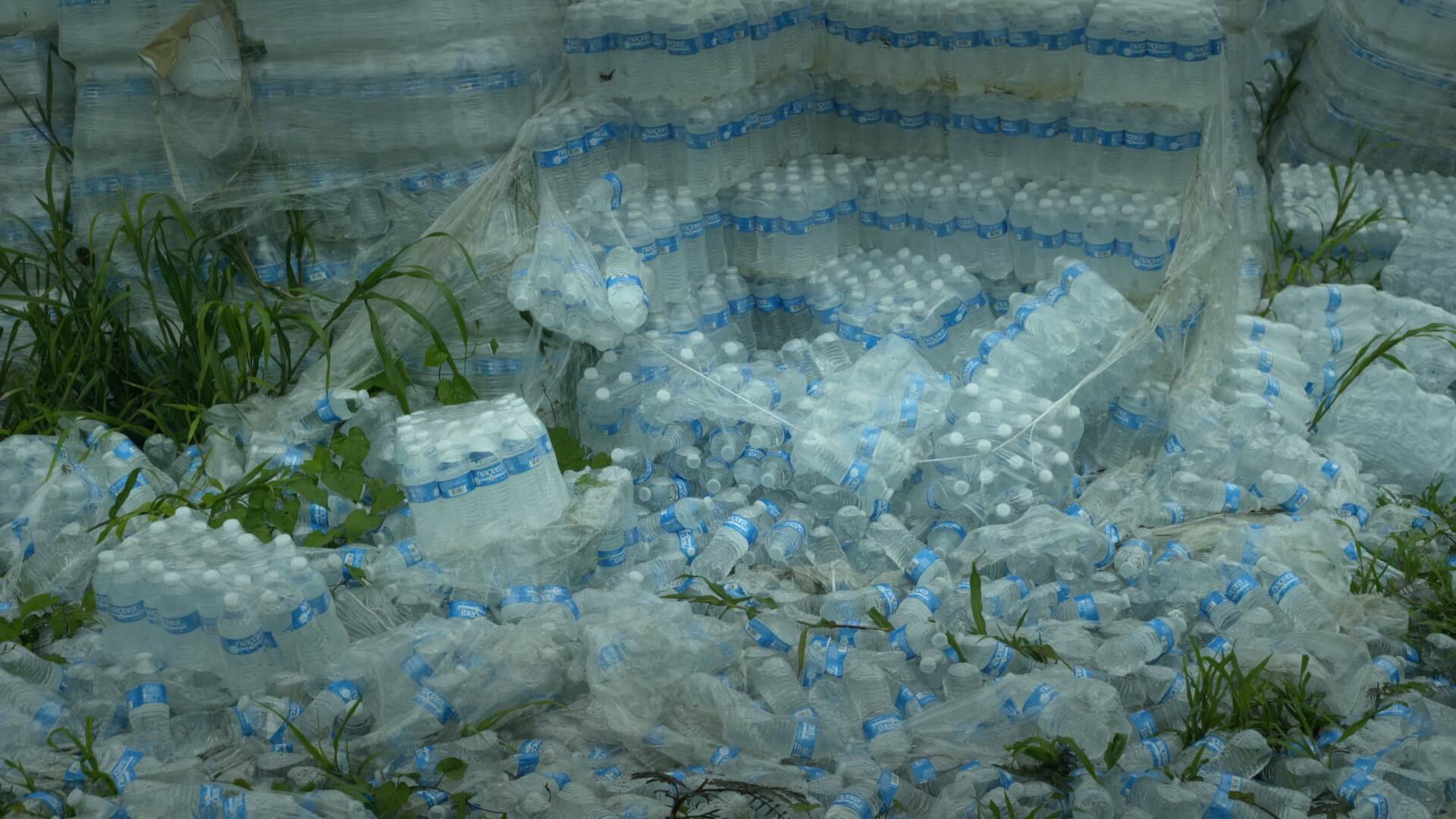 Still from Landfall. Hundreds of plastic bottles are packaged in plastic. Some of the plastic is broken open and the bottles have been spread all over the ground.