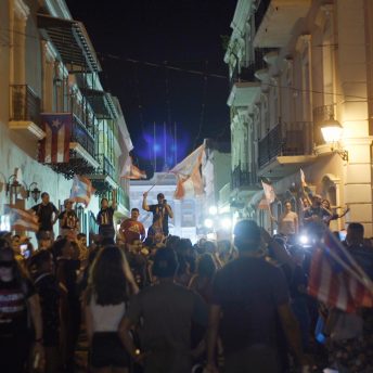 Still from Landfall. It is nighttime and in the middle of a street there is a crowd of people with flags and t-shirts with the Puerto Rican flag.
