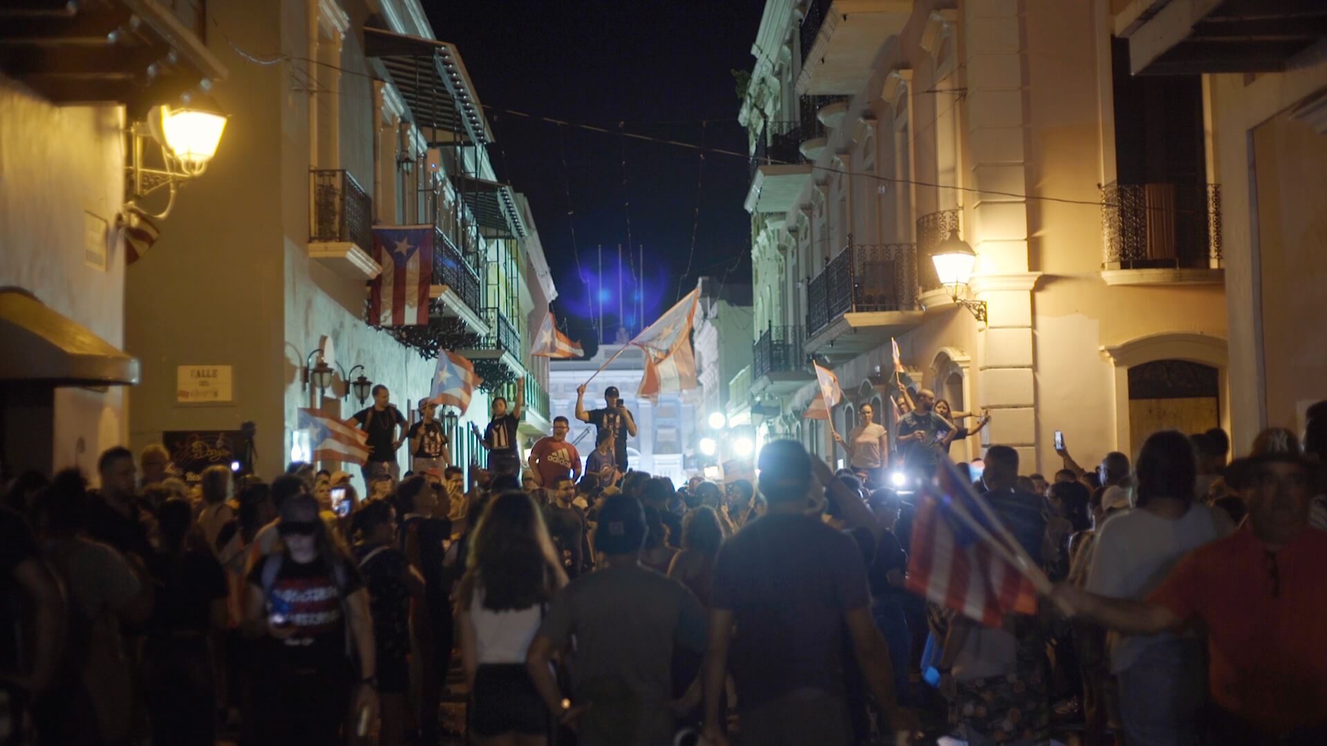 Still from Landfall. It is nighttime and in the middle of a street there is a crowd of people with flags and t-shirts with the Puerto Rican flag.