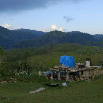 Still from Landfall. Full shot of a green landscape with mountains in the background. On the right, there is a house with a wooden pillar front porch and a pickup truck is parked to the side of the house.