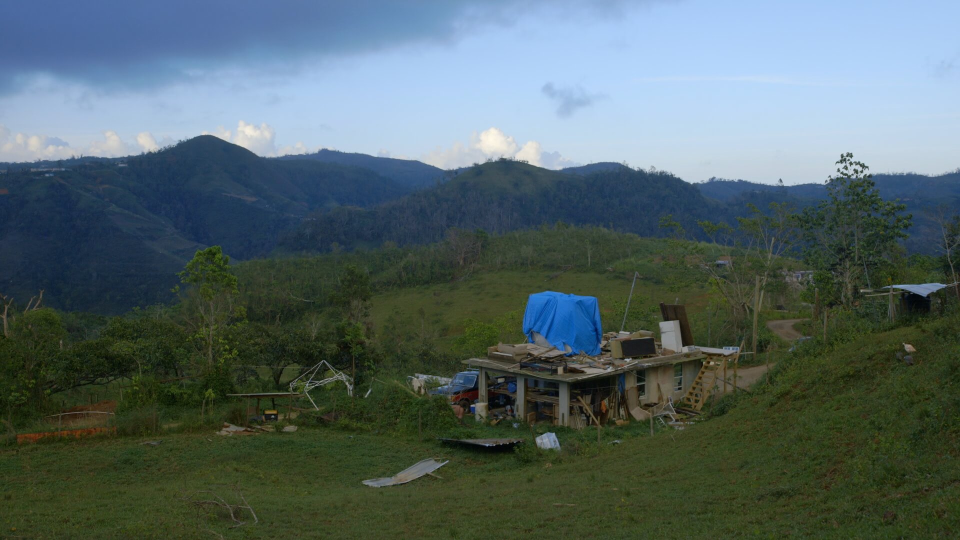 Still from Landfall. Full shot of a green landscape with mountains in the background. On the right, there is a house with a wooden pillar front porch and a pickup truck is parked to the side of the house.