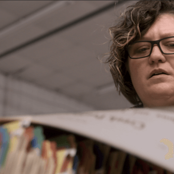 Angel Ellis is an Indigenous woman wearing dark glasses. She has blonde curly hair and a concerned look as she reads newspapers from the physical archives in the Mvskoke Media warehouse.