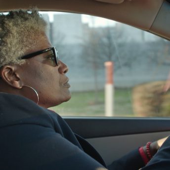 Still from I am not going to change 400 years in four. Medium shot of a woman driving a car, she has black thick glasses, short curly white hair, and wears a black hoodie and silver hoop earrings.