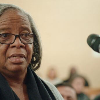 Still from I am not going to change 400 years in four. Close-up of a woman talking in front of a microphone. She has medium-length gray hair, and wears a black scarf and a gray shirt.