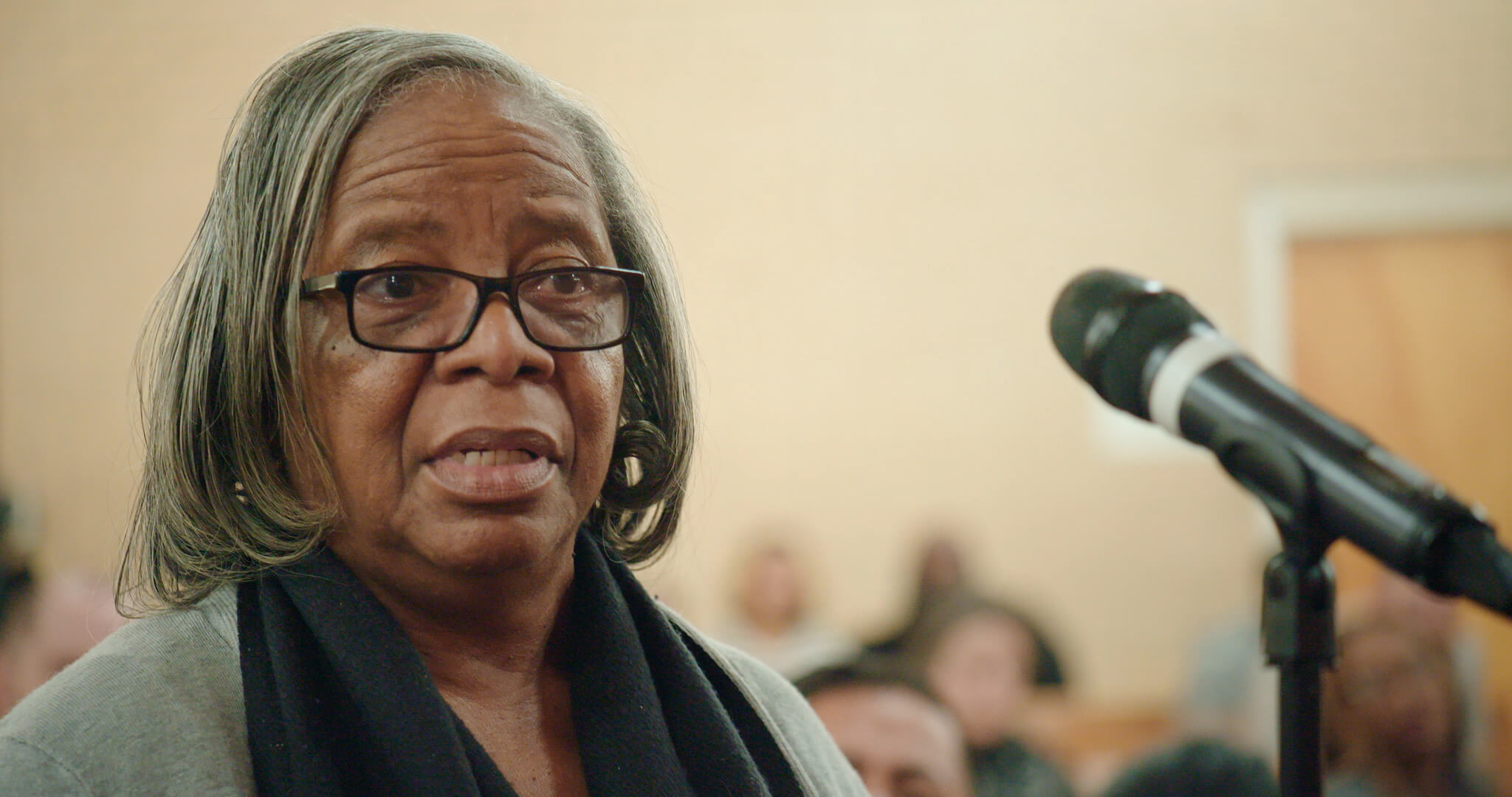 Still from I am not going to change 400 years in four. Close-up of a woman talking in front of a microphone. She has medium-length gray hair, and wears a black scarf and a gray shirt.