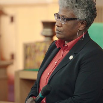 Still from I am not going to change 400 years in four. Medium shot of a woman wearing a red shirt, black blazer, and thick glasses. She has curly short white hair. She holds a microphone and looks at something outside of the shot.