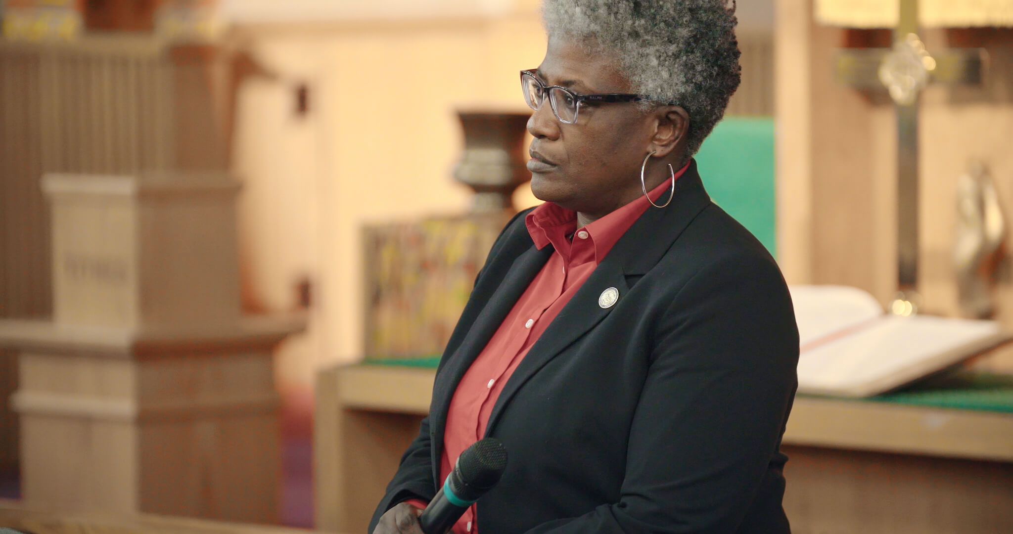 Still from I am not going to change 400 years in four. Medium shot of a woman wearing a red shirt, black blazer, and thick glasses. She has curly short white hair. She holds a microphone and looks at something outside of the shot.