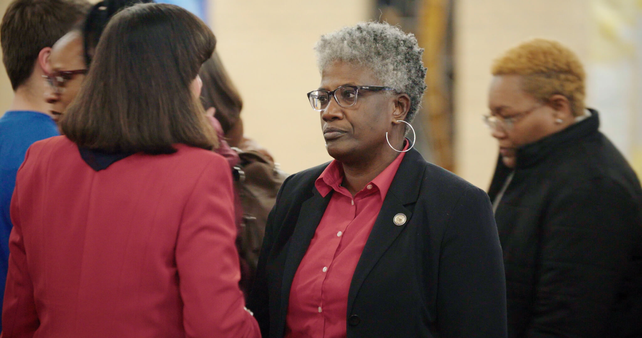 Still from I am not going to change 400 years in four. Two women talking to each other, one wears a red blazer, and the other one has short hair, a red shirt, a black blazer, and glasses.